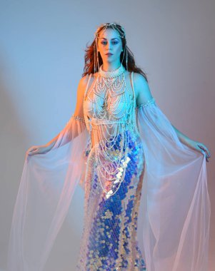 Peri masalı kostümü giymiş kızıl saçlı bir deniz kızı perisinin fantezi stüdyosu portresi. Akan kumaş ve ruhani stüdyo ışıklandırmasıyla klasik yürüyüş pozu, izole edilmiş arka plan.