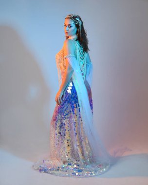 Peri masalı kostümü giymiş kızıl saçlı bir deniz kızı perisinin fantezi stüdyosu portresi. Akan kumaş ve ruhani stüdyo ışıklandırmasıyla klasik yürüyüş pozu, izole edilmiş arka plan.