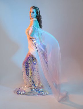 Peri masalı kostümü giymiş kızıl saçlı bir deniz kızı perisinin fantezi stüdyosu portresi. Akan kumaş ve ruhani stüdyo ışıklandırmasıyla klasik yürüyüş pozu, izole edilmiş arka plan.