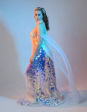 Peri masalı kostümü giymiş kızıl saçlı bir deniz kızı perisinin fantezi stüdyosu portresi. Akan kumaş ve ruhani stüdyo ışıklandırmasıyla klasik yürüyüş pozu, izole edilmiş arka plan.