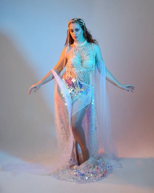 Peri masalı kostümü giymiş kızıl saçlı bir deniz kızı perisinin fantezi stüdyosu portresi. Akan kumaş ve ruhani stüdyo ışıklandırmasıyla klasik yürüyüş pozu, izole edilmiş arka plan.