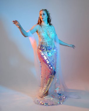 Peri masalı kostümü giymiş kızıl saçlı bir deniz kızı perisinin fantezi stüdyosu portresi. Akan kumaş ve ruhani stüdyo ışıklandırmasıyla klasik yürüyüş pozu, izole edilmiş arka plan.