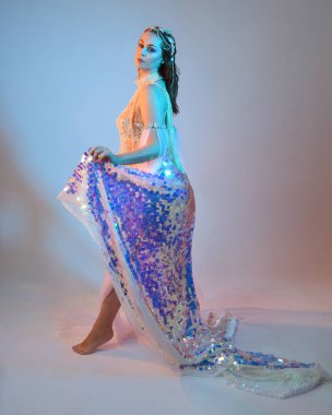 Peri masalı kostümü giymiş kızıl saçlı bir deniz kızı perisinin fantezi stüdyosu portresi. Akan kumaş ve ruhani stüdyo ışıklandırmasıyla klasik yürüyüş pozu, izole edilmiş arka plan.