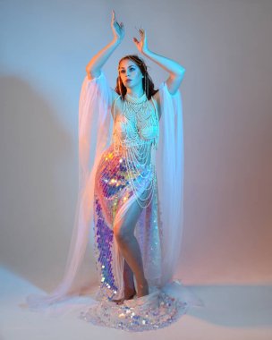 Peri masalı kostümü giymiş kızıl saçlı bir deniz kızı perisinin fantezi stüdyosu portresi. Akan kumaş ve ruhani stüdyo ışıklandırmasıyla klasik yürüyüş pozu, izole edilmiş arka plan.