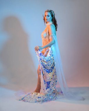 Peri masalı kostümü giymiş kızıl saçlı bir deniz kızı perisinin fantezi stüdyosu portresi. Akan kumaş ve ruhani stüdyo ışıklandırmasıyla klasik yürüyüş pozu, izole edilmiş arka plan.