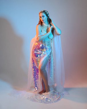 Peri masalı kostümü giymiş kızıl saçlı bir deniz kızı perisinin fantezi stüdyosu portresi. Akan kumaş ve ruhani stüdyo ışıklandırmasıyla klasik yürüyüş pozu, izole edilmiş arka plan.