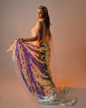 Peri masalı kostümü giymiş kızıl saçlı bir deniz kızı perisinin fantezi stüdyosu portresi. Akan kumaş ve ruhani stüdyo ışıklandırmasıyla klasik yürüyüş pozu, izole edilmiş arka plan.