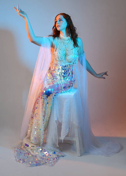 Fantasy studio portrait of a red-haired mermaid faery model wearing a white pearl gown, fairytale costume. classical sitting pose with flowing fabric and ethereal studio lighting, isolated background.