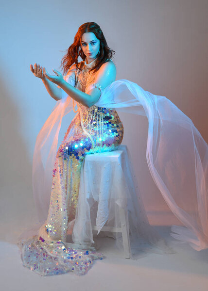 Fantasy studio portrait of a red-haired mermaid faery model wearing a white pearl gown, fairytale costume. classical sitting pose with flowing fabric and ethereal studio lighting, isolated background.