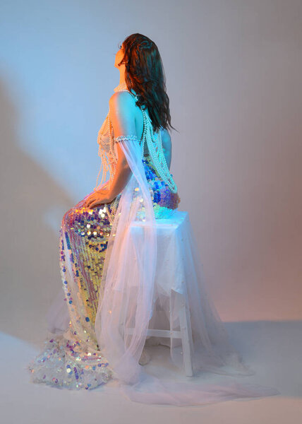Fantasy studio portrait of a red-haired mermaid faery model wearing a white pearl gown, fairytale costume. classical sitting pose with flowing fabric and ethereal studio lighting, isolated background.