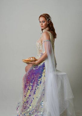 Fantezi stüdyo portresi, beyaz inci elbise giyen kızıl saçlı bir deniz kızı peri modeli, peri masalı kostümü. Klasik oturma pozu deniz kabuğu, stüdyo arka planında ruhani aydınlatma.