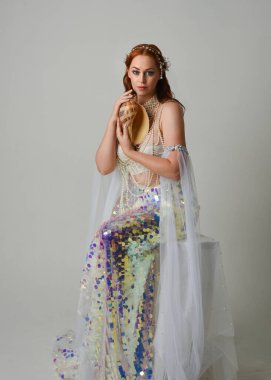 Fantezi stüdyo portresi, beyaz inci elbise giyen kızıl saçlı bir deniz kızı peri modeli, peri masalı kostümü. Klasik oturma pozu deniz kabuğu, stüdyo arka planında ruhani aydınlatma.