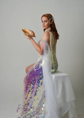 Fantezi stüdyo portresi, beyaz inci elbise giyen kızıl saçlı bir deniz kızı peri modeli, peri masalı kostümü. Klasik oturma pozu deniz kabuğu, stüdyo arka planında ruhani aydınlatma.