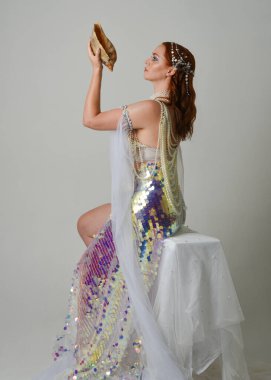 Fantezi stüdyo portresi, beyaz inci elbise giyen kızıl saçlı bir deniz kızı peri modeli, peri masalı kostümü. Klasik oturma pozu deniz kabuğu, stüdyo arka planında ruhani aydınlatma.