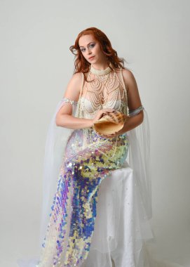 Fantezi stüdyo portresi, beyaz inci elbise giyen kızıl saçlı bir deniz kızı peri modeli, peri masalı kostümü. Klasik oturma pozu deniz kabuğu, stüdyo arka planında ruhani aydınlatma.