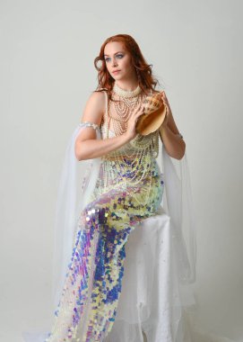 Fantezi stüdyo portresi, beyaz inci elbise giyen kızıl saçlı bir deniz kızı peri modeli, peri masalı kostümü. Klasik oturma pozu deniz kabuğu, stüdyo arka planında ruhani aydınlatma.