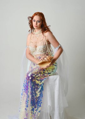 Fantezi stüdyo portresi, beyaz inci elbise giyen kızıl saçlı bir deniz kızı peri modeli, peri masalı kostümü. Klasik oturma pozu deniz kabuğu, stüdyo arka planında ruhani aydınlatma.