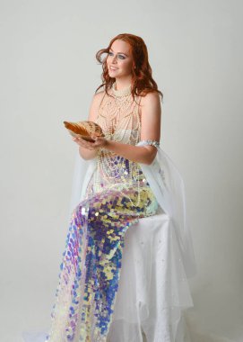 Fantezi stüdyo portresi, beyaz inci elbise giyen kızıl saçlı bir deniz kızı peri modeli, peri masalı kostümü. Klasik oturma pozu deniz kabuğu, stüdyo arka planında ruhani aydınlatma.