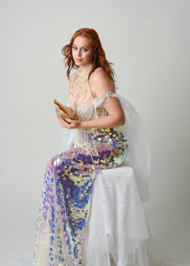 Fantezi stüdyo portresi, beyaz inci elbise giyen kızıl saçlı bir deniz kızı peri modeli, peri masalı kostümü. Klasik oturma pozu deniz kabuğu, stüdyo arka planında ruhani aydınlatma.