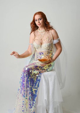 Fantezi stüdyo portresi, beyaz inci elbise giyen kızıl saçlı bir deniz kızı peri modeli, peri masalı kostümü. Klasik oturma pozu deniz kabuğu, stüdyo arka planında ruhani aydınlatma.