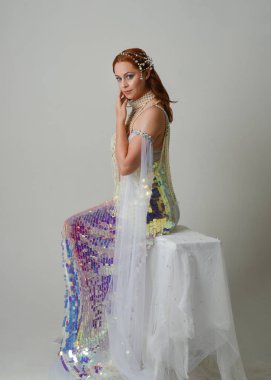 Fantezi stüdyo portresi, beyaz inci elbise giyen kızıl saçlı bir deniz kızı peri modeli, peri masalı kostümü. Klasik oturma pozu ters dönük, stüdyo arka planında ruhani aydınlatma.