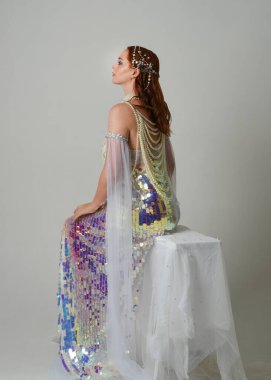 Fantezi stüdyo portresi, beyaz inci elbise giyen kızıl saçlı bir deniz kızı peri modeli, peri masalı kostümü. Klasik oturma pozu ters dönük, stüdyo arka planında ruhani aydınlatma.