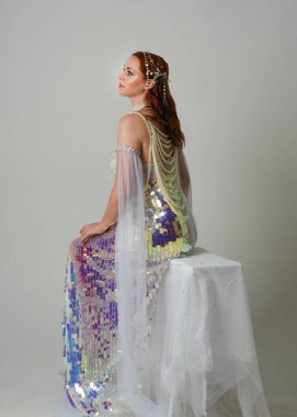 Fantezi stüdyo portresi, beyaz inci elbise giyen kızıl saçlı bir deniz kızı peri modeli, peri masalı kostümü. Klasik oturma pozu ters dönük, stüdyo arka planında ruhani aydınlatma.