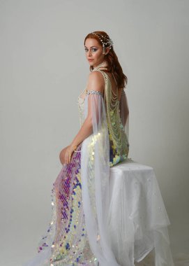 Fantezi stüdyo portresi, beyaz inci elbise giyen kızıl saçlı bir deniz kızı peri modeli, peri masalı kostümü. Klasik oturma pozu ters dönük, stüdyo arka planında ruhani aydınlatma.