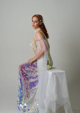 Fantezi stüdyo portresi, beyaz inci elbise giyen kızıl saçlı bir deniz kızı peri modeli, peri masalı kostümü. Klasik oturma pozu ters dönük, stüdyo arka planında ruhani aydınlatma.