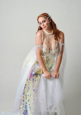 Fantezi stüdyo portresi, beyaz inci elbise giyen kızıl saçlı bir deniz kızı peri modeli, peri masalı kostümü. Klasik oturma pozu ters dönük, stüdyo arka planında ruhani aydınlatma.