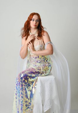 Fantezi stüdyo portresi, beyaz inci elbise giyen kızıl saçlı bir deniz kızı peri modeli, peri masalı kostümü. Klasik oturma pozu ters dönük, stüdyo arka planında ruhani aydınlatma.