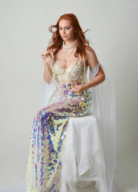 Fantezi stüdyo portresi, beyaz inci elbise giyen kızıl saçlı bir deniz kızı peri modeli, peri masalı kostümü. Klasik oturma pozu ters dönük, stüdyo arka planında ruhani aydınlatma.