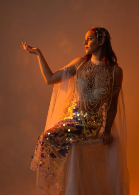 Fantezi stüdyo portresi, beyaz inci elbise giyen kızıl saçlı bir deniz kızı peri modeli, peri masalı kostümü. Klasik oturma pozu ters dönük, stüdyo arka planında ruhani aydınlatma.