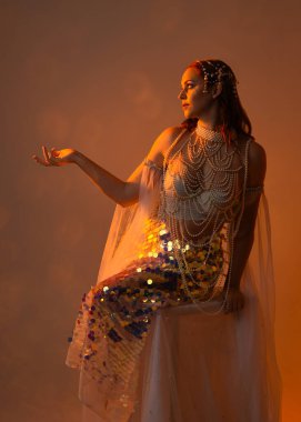 Fantezi stüdyo portresi, beyaz inci elbise giyen kızıl saçlı bir deniz kızı peri modeli, peri masalı kostümü. Klasik oturma pozu ters dönük, stüdyo arka planında ruhani aydınlatma.