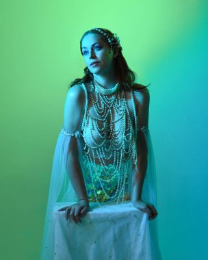 Fantezi stüdyo portresi, beyaz inci elbise giyen kızıl saçlı bir deniz kızı peri modeli, peri masalı kostümü. Klasik oturma pozu, ruhsal karamsar ışıklandırma stüdyo arka planında su altındaymış gibi.