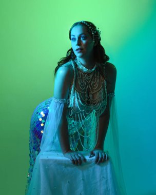 Fantezi stüdyo portresi, beyaz inci elbise giyen kızıl saçlı bir deniz kızı peri modeli, peri masalı kostümü. Klasik oturma pozu, ruhsal karamsar ışıklandırma stüdyo arka planında su altındaymış gibi.