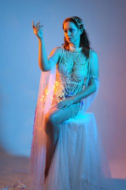 Fantezi stüdyo portresi, beyaz inci elbise giyen kızıl saçlı bir deniz kızı peri modeli, peri masalı kostümü. Klasik oturma pozu, ruhsal karamsar ışıklandırma stüdyo arka planında su altındaymış gibi.