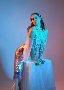 Fantezi stüdyo portresi, beyaz inci elbise giyen kızıl saçlı bir deniz kızı peri modeli, peri masalı kostümü. Klasik oturma pozu, ruhsal karamsar ışıklandırma stüdyo arka planında su altındaymış gibi.