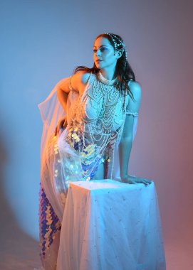 Fantezi stüdyo portresi, beyaz inci elbise giyen kızıl saçlı bir deniz kızı peri modeli, peri masalı kostümü. Klasik oturma pozu, ruhsal karamsar ışıklandırma stüdyo arka planında su altındaymış gibi.