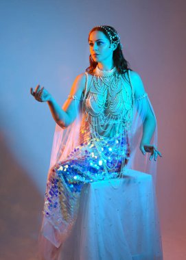 Fantezi stüdyo portresi, beyaz inci elbise giyen kızıl saçlı bir deniz kızı peri modeli, peri masalı kostümü. Klasik oturma pozu, ruhsal karamsar ışıklandırma stüdyo arka planında su altındaymış gibi.