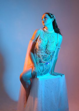 Fantezi stüdyo portresi, beyaz inci elbise giyen kızıl saçlı bir deniz kızı peri modeli, peri masalı kostümü. Klasik oturma pozu, ruhsal karamsar ışıklandırma stüdyo arka planında su altındaymış gibi.