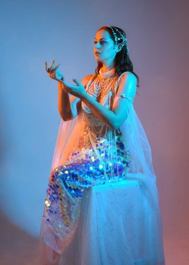 Fantezi stüdyo portresi, beyaz inci elbise giyen kızıl saçlı bir deniz kızı peri modeli, peri masalı kostümü. Klasik oturma pozu, ruhsal karamsar ışıklandırma stüdyo arka planında su altındaymış gibi.