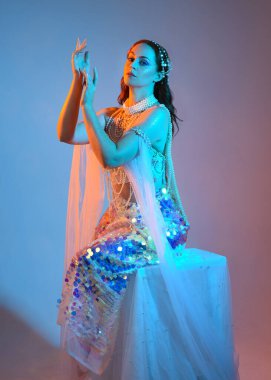 Fantezi stüdyo portresi, beyaz inci elbise giyen kızıl saçlı bir deniz kızı peri modeli, peri masalı kostümü. Klasik oturma pozu, ruhsal karamsar ışıklandırma stüdyo arka planında su altındaymış gibi.