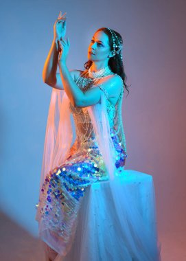 Fantezi stüdyo portresi, beyaz inci elbise giyen kızıl saçlı bir deniz kızı peri modeli, peri masalı kostümü. Klasik oturma pozu, ruhsal karamsar ışıklandırma stüdyo arka planında su altındaymış gibi.