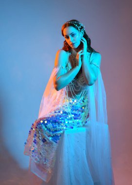 Fantezi stüdyo portresi, beyaz inci elbise giyen kızıl saçlı bir deniz kızı peri modeli, peri masalı kostümü. Klasik oturma pozu, ruhsal karamsar ışıklandırma stüdyo arka planında su altındaymış gibi.