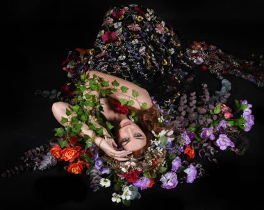 Celtic Bahar Tanrıçası Olarak Kadın Model Stüdyo Portreleri, Beltane Pagan Mitolojisinden esinlenerek Çiçek Fantezi Pelerini Kostümü. Klasik oturma pozu çiçek yatağında, izole edilmiş Karanlık Arkaplan