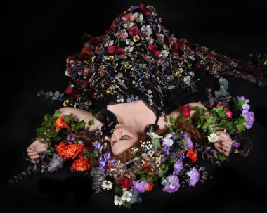 Celtic Bahar Tanrıçası Olarak Kadın Model Stüdyo Portreleri, Beltane Pagan Mitolojisinden esinlenerek Çiçek Fantezi Pelerini Kostümü. Klasik oturma pozu çiçek yatağında, izole edilmiş Karanlık Arkaplan