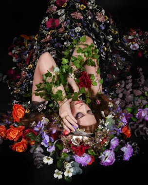 Celtic Bahar Tanrıçası Olarak Kadın Model Stüdyo Portreleri, Beltane Pagan Mitolojisinden esinlenerek Çiçek Fantezi Pelerini Kostümü. Klasik oturma pozu çiçek yatağında, izole edilmiş Karanlık Arkaplan