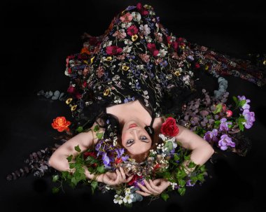 Celtic Bahar Tanrıçası Olarak Kadın Model Stüdyo Portreleri, Beltane Pagan Mitolojisinden esinlenerek Çiçek Fantezi Pelerini Kostümü. Klasik oturma pozu çiçek yatağında, izole edilmiş Karanlık Arkaplan