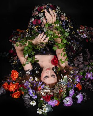 Celtic Bahar Tanrıçası Olarak Kadın Model Stüdyo Portreleri, Beltane Pagan Mitolojisinden esinlenerek Çiçek Fantezi Pelerini Kostümü. Klasik oturma pozu çiçek yatağında, izole edilmiş Karanlık Arkaplan